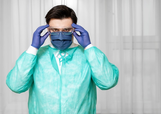 Photo tired doctor in protective equipment at home puts on protective medical mask with glasses, coronavirus epidemic