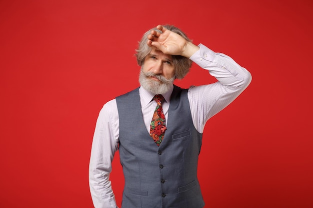 Tired elderly gray-haired mustache bearded man in classic shirt vest and colorful tie isolated on red wall background in studio. People lifestyle concept. Mock up copy space. Putting hand on forehead.