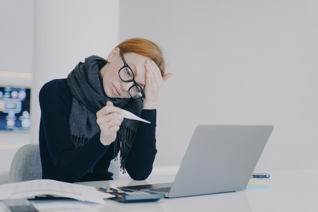 Tired executive caught cold and measuring temperature Sad young white woman in scarf is sick