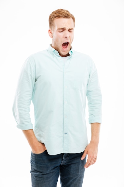 Tired exhausted young man standing and yawning over white wall