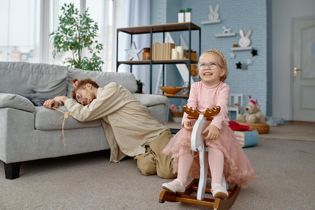 Tired fatigue father sleeping on floor and cute naughty girl playing at home