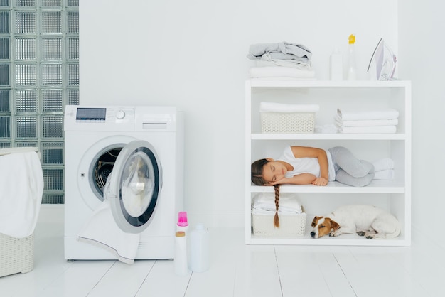 Tired female child rests on console shelf together with pet does washing at home sleeps in laundry room opened washing machine with dirty towel inside Childhood cleanliness family chores concept