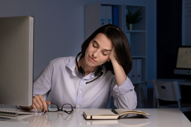 Tired female hotline operator with closed eyes napping by desk while working late in the evening in dark office