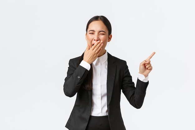 Tired female office manager, entrepreneur in suit pointing finger right and yawning. Bored or exhausted asian businesswoman working late hours, showing project, standing white background