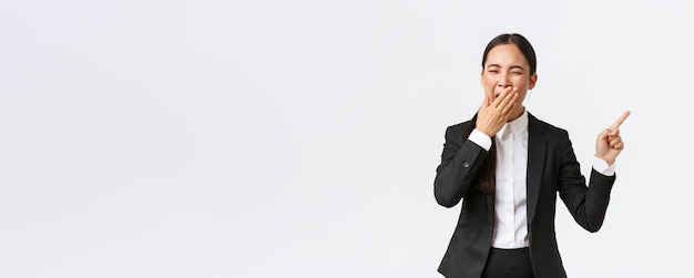 Tired female office manager entrepreneur in suit pointing finger right and yawning Bored or exhausted asian businesswoman working late hours showing project standing white background