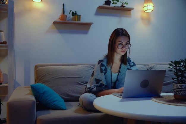 Tired focused woman using laptop at night working overtime at home sitting on sofa flexible working hours