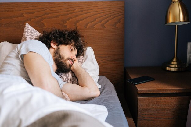 Tired handsome bearded young man sleeping with open eyes peacefully lying on back in large comfortable double bed under white blanket