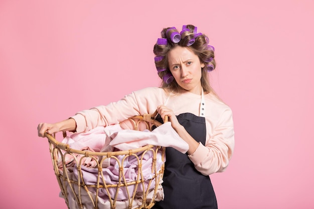 Tired of household chores the woman holds a wicker basket with laundry clothes posing against a pink background The girl has a sad face does not like cleaning