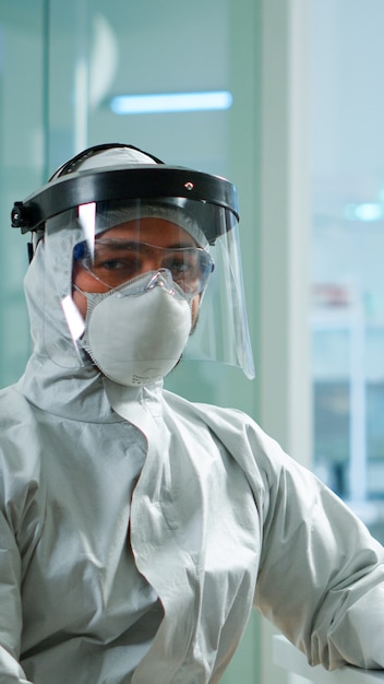 Tired laboratory doctor with overall suit working at computer and looking at camera. Scientist examining virus evolution using high tech, chemistry tools for scientific research, vaccine development