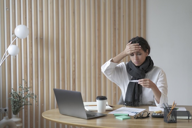 Tired lady freelancer sitting at desk at home office looking sickly at digital thermometer