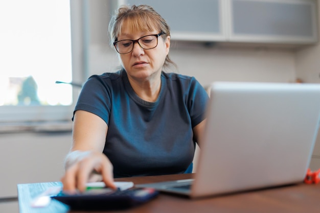 Tired mature blond woman freelancer working from home using laptop sitting in kitchen