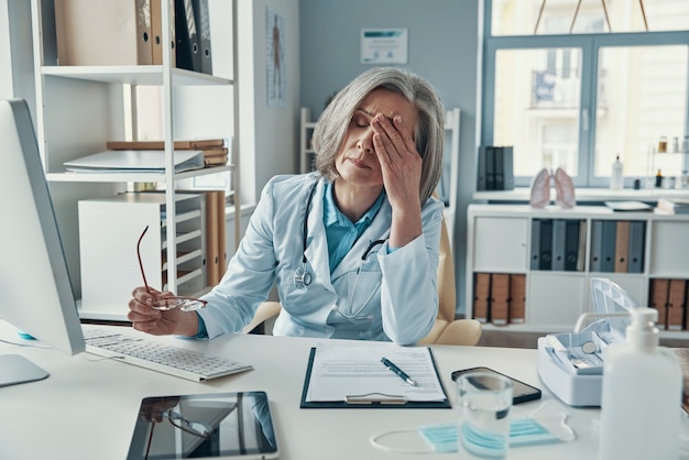 Tired mature female doctor in white lab coat covering her face with hand while sitting in her office