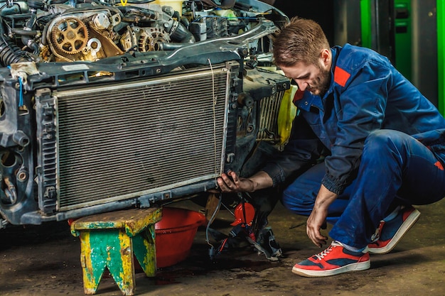 A tired mechanic in a blue protective suit is repairing a car radiator. Repair Service Concept. 