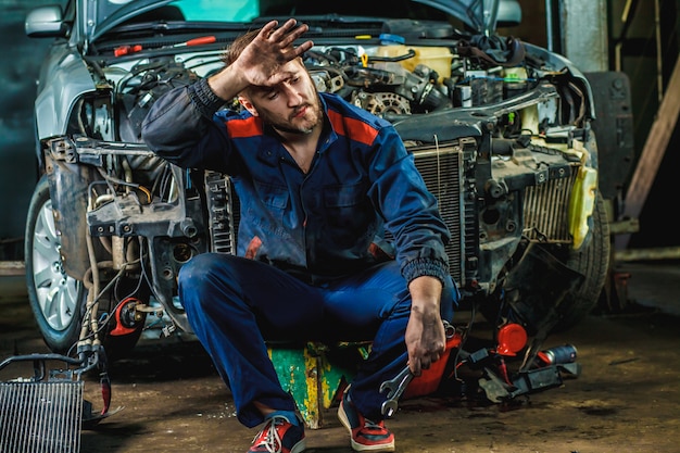 A tired mechanic in a blue protective suit is sitting near a disassembled car.