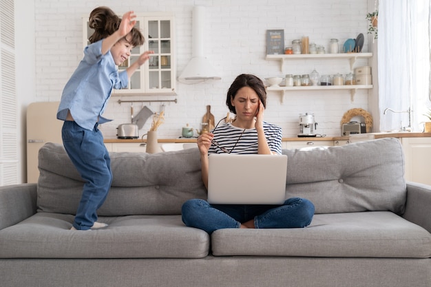 Tired mother sit on couch, work on laptop at home, hyperactive little kid jumping drawing attention.