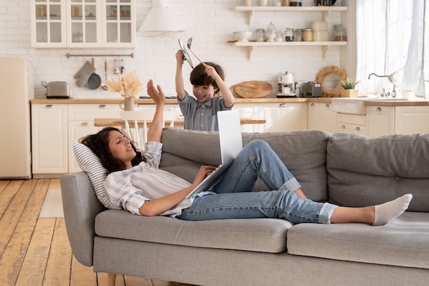 Tired mother try to concentrate on work type business email on laptop on couch with active noisy kid