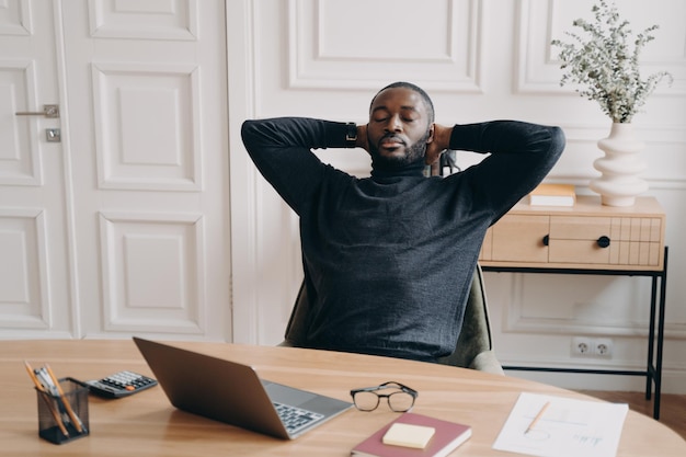 Tired overworked african american male office worker resting with eyes closed and hands behind head