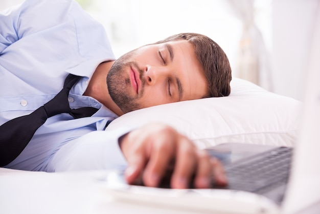 Tired and overworked. Handsome young man in shirt and tie sleeping in bed with his hand laying on laptop keyboard