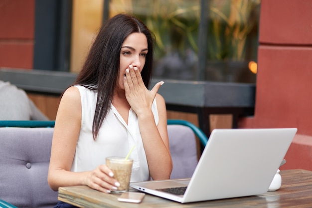 Tired overworked young female freelnacer yawns as want to sleep, wants to have rest, works long time at laptop computer, drinks cocktai