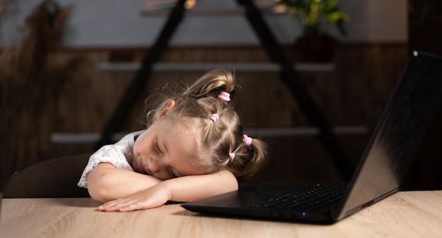 Tired preschool girl sits at the table and sleeps near the laptop