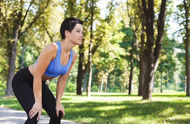 Tired runner breathing, taking run break in park. Athlete woman with hands on knees having rest after workout, copy space