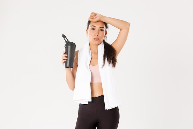 Tired but satisfied asian fitness girl wiping sweat from forehead and exhale after good workout, drinking water or protein during training session.