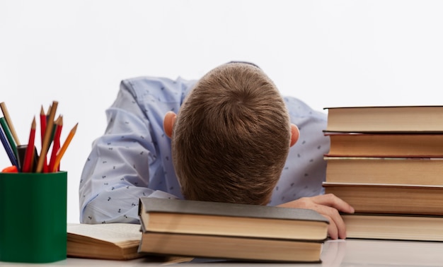 tired schoolboy sits at a table and rested his head on textbooks.