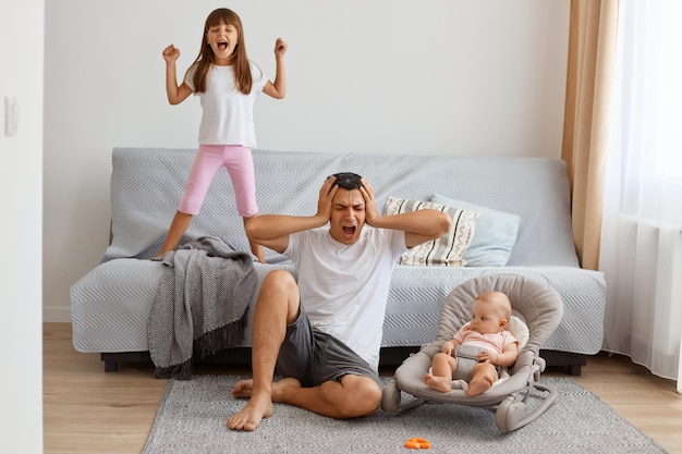 Tired screaming handsome brunette father spending time with daughters while sitting on floor near sofa, covering ears with hands, wants not to hear the screaming older daughter.