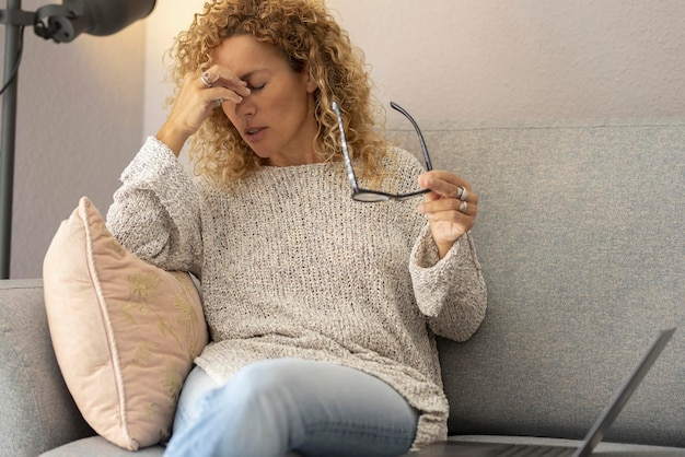 Tired stressed adult woman with headache touch his nose and front sitting on the sofa. Unhealthy lifestyle and home problems concept people. Female with flu illness firsts symptoms