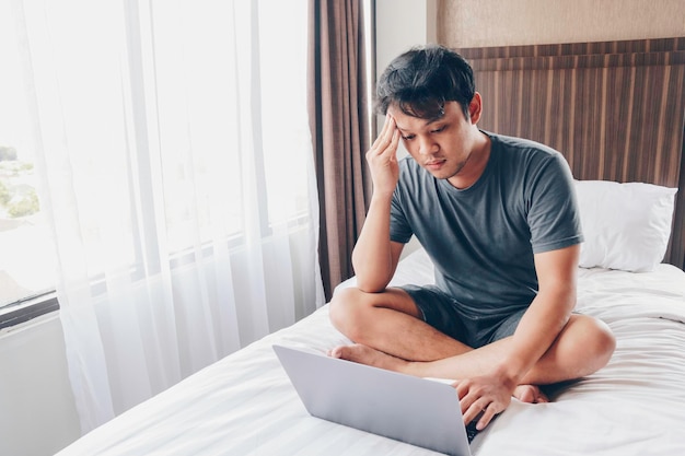 Tired stressed young Asian man feeling sleepy and tired while using laptop on the bed in bedroom Hard work concept