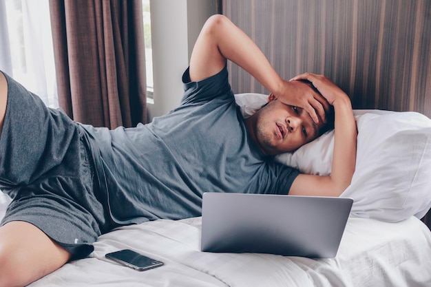 Tired stressed young Asian man feeling sleepy and tired while using laptop on the bed in bedroom Hard work concept