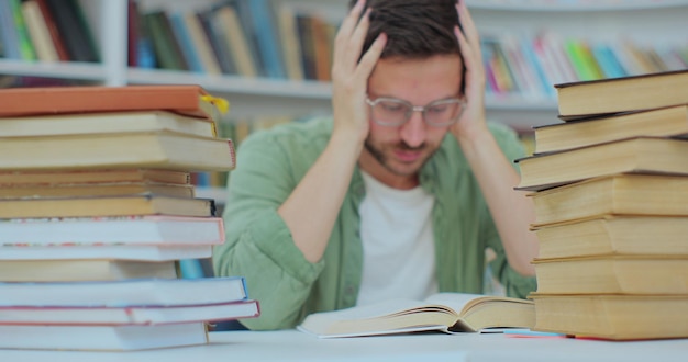 A tired student sits at desk full of books Young man preparing for an exam in the college library Self education concept