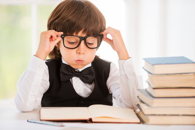 Tired of studying. Tired little boy looking at the book and holding his glasses while sitting at the table