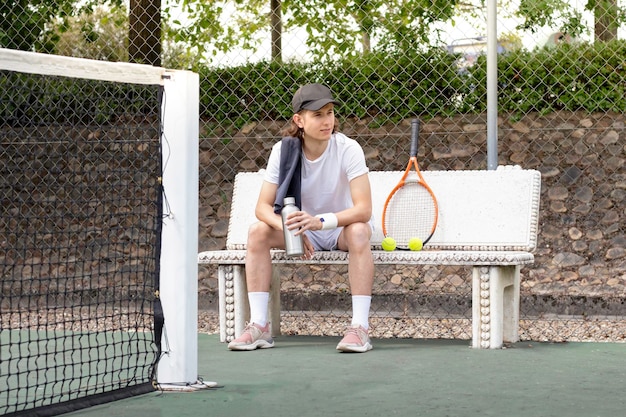 Tired tennis player man looking sideways while sitting on a bench on a tennis court with cap on and a towel