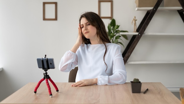 Tired tutor girl with headache sits at a desk at home giving an online lecture Woman blogger broadcasts live on smartphone camera Distance education and work Blogging