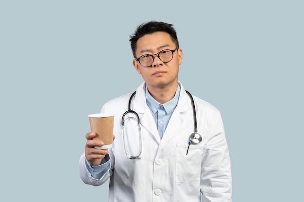 Tired unhappy middle aged chinese male doctor in white coat and glasses with cup of coffee takeaway