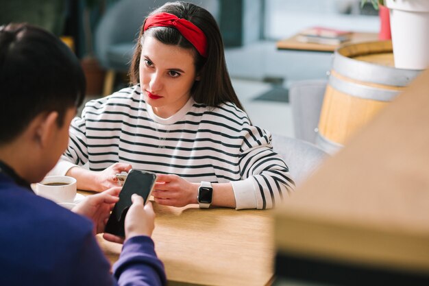 Tired unsatisfied lady looking attentively at the man sitting with her while holding a smartphone and watching him using his gadget