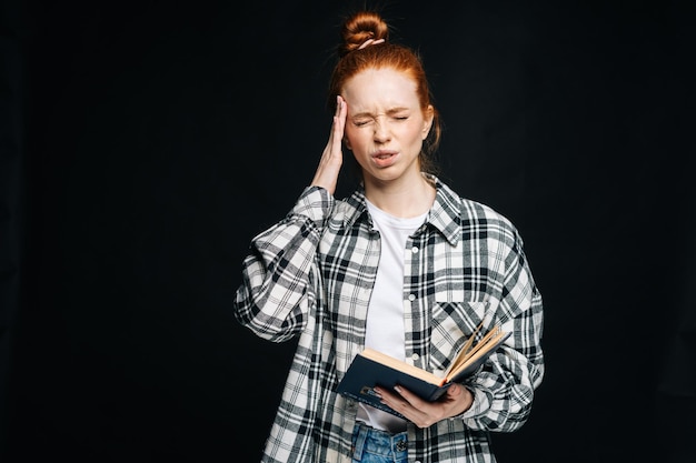 Tired upset young woman college student with headache holding book and touching temples on isolated black background Pretty redhead lady model emotionally showing facial expressions in studio