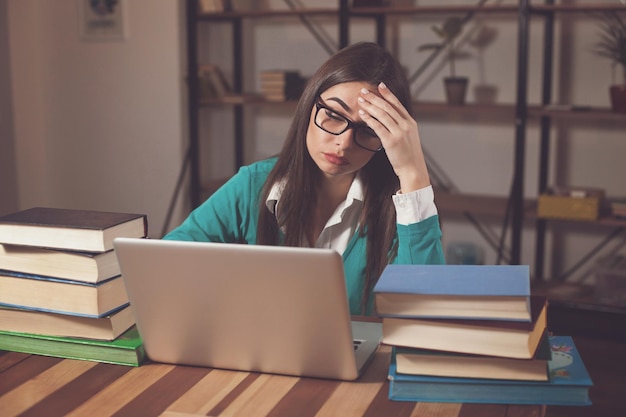 Tired woman in glasses with lots of books and laptop is sitting at the table