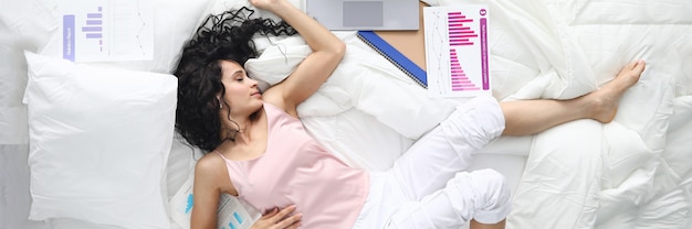 Tired woman in pajamas sleep in pose in white bed. There is laptop on bed, scattered document with text and graphic, and notebook.