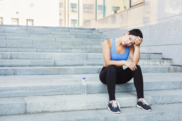 Tired woman runner is having break, sitting on steps outdoors. City jogging and active lifestyle concept, copy space