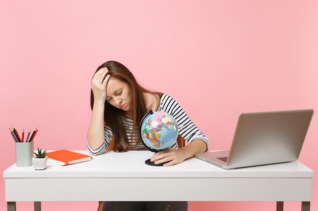 Tired woman with closed eyes lean on hand hold globe having problems with vacation planning while sit, work at office with laptop isolated on pink background. Achievement business career. Copy space.