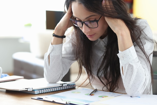 Tired woman with glasses sit at table and massages her head with her hands and think