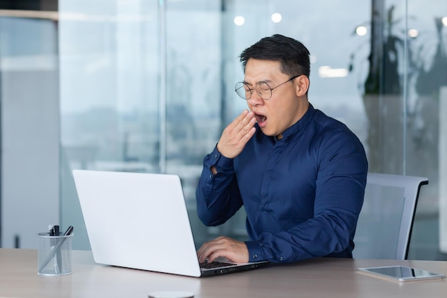 Tired worker at work yawning asian man in shirt and glasses working inside office using laptop man