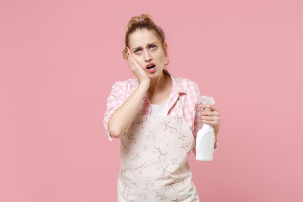 Tired worried young woman housewife in casual clothes, apron hold atomizer spray with washing cleanser doing housework isolated on pink background in studio. Housekeeping concept. Put hand on cheek.