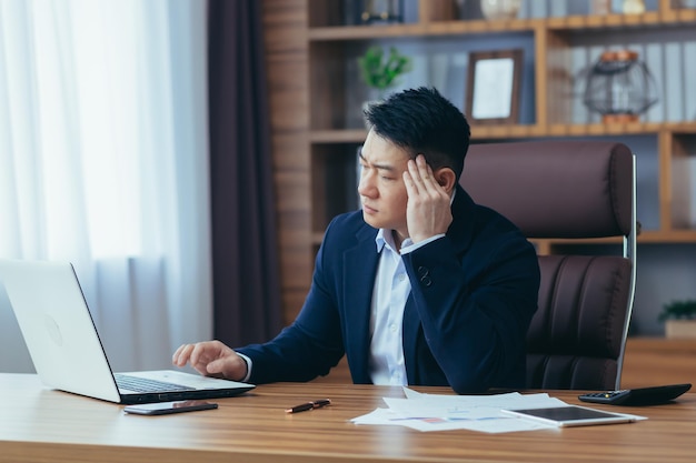 Tired young asian man businessman worker lawyer at work He has a headache keeps his hands on his head sits at a desk in the office