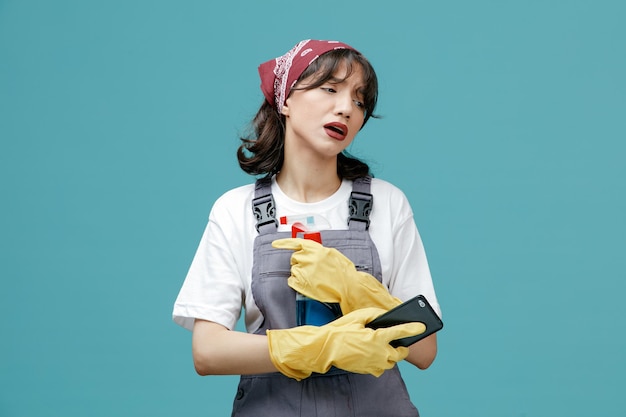 Tired young female cleaner wearing uniform bandana and rubber gloves holding cleanser and mobile phone looking at side isolated on blue background