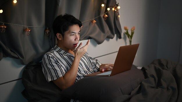 Tired young man working with laptop computer and in bedroom at night.