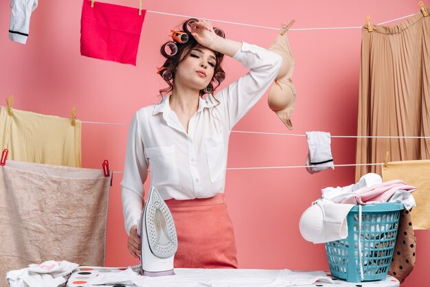 Tired young woman housewife in shirt, clothes on rope and ironing clean clothes on board doing housework isolated on pink wall