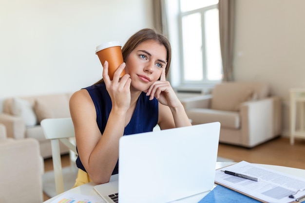 Tired young woman looking away and holding cup of coffee near head in office on blurred foreground